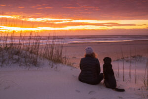 Woman and dog watch sunset while sitting on sanding dune of a North Carolina beach enjoying her co-ownership vacation home